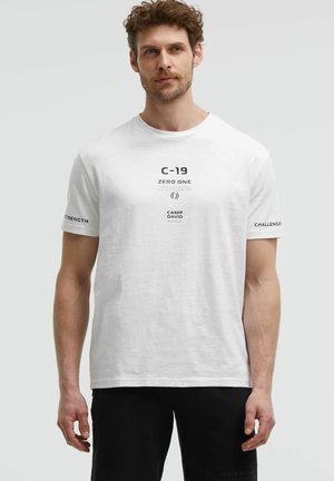 Man wearing white t-shirt with text "C-19 Zero One Camp David Workout" and black shorts, standing against plain light background.