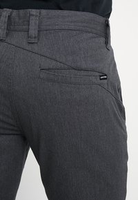 Gros plan d'une personne portant un pantalon gris foncé avec une couture diagonale et une petite étiquette VOLCOM au-dessus de la poche arrière.