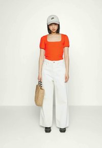 Top curto de mangas curtas em laranja brilhante com nervuras, jeans brancos de perna larga, boné cinza da Nike, mala de cesto com alças pretas, sapatos pretos de plataforma.