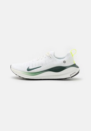 Weißer Sportschuh mit grünem Swoosh-Logo, dicker grüner und weißer Sohle und gelbem Zuglasche an der Ferse, seitlich auf neutralem Hintergrund gezeigt.
