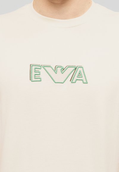 Κοντινό πλάνο ενός λευκού t-shirt με πράσινο και μπεζ κεντητό λογότυπο που γράφει «EVA» στο ύψος του στήθους.