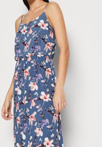 Robe maxi florale avec un fond bleu, ornée de fleurs roses et violettes ainsi que de papillons, bretelles spaghetti ajustables et taille froncée.