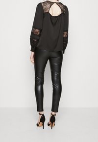 Svart blus med spetsdetaljer och nyckelhål bak, som matchas med svarta leggings i syntetläder. Modellen bär svarta högklackade skor.