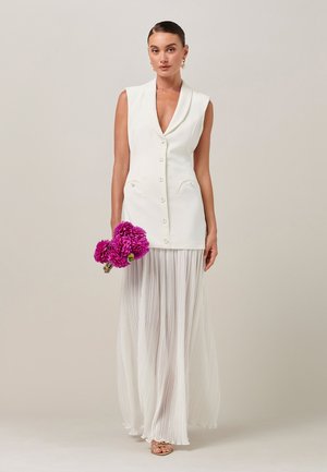Femme portant un gilet blanc sans manches boutonné et une longue jupe plissée, tenant des fleurs roses vives, debout devant un fond clair uni.