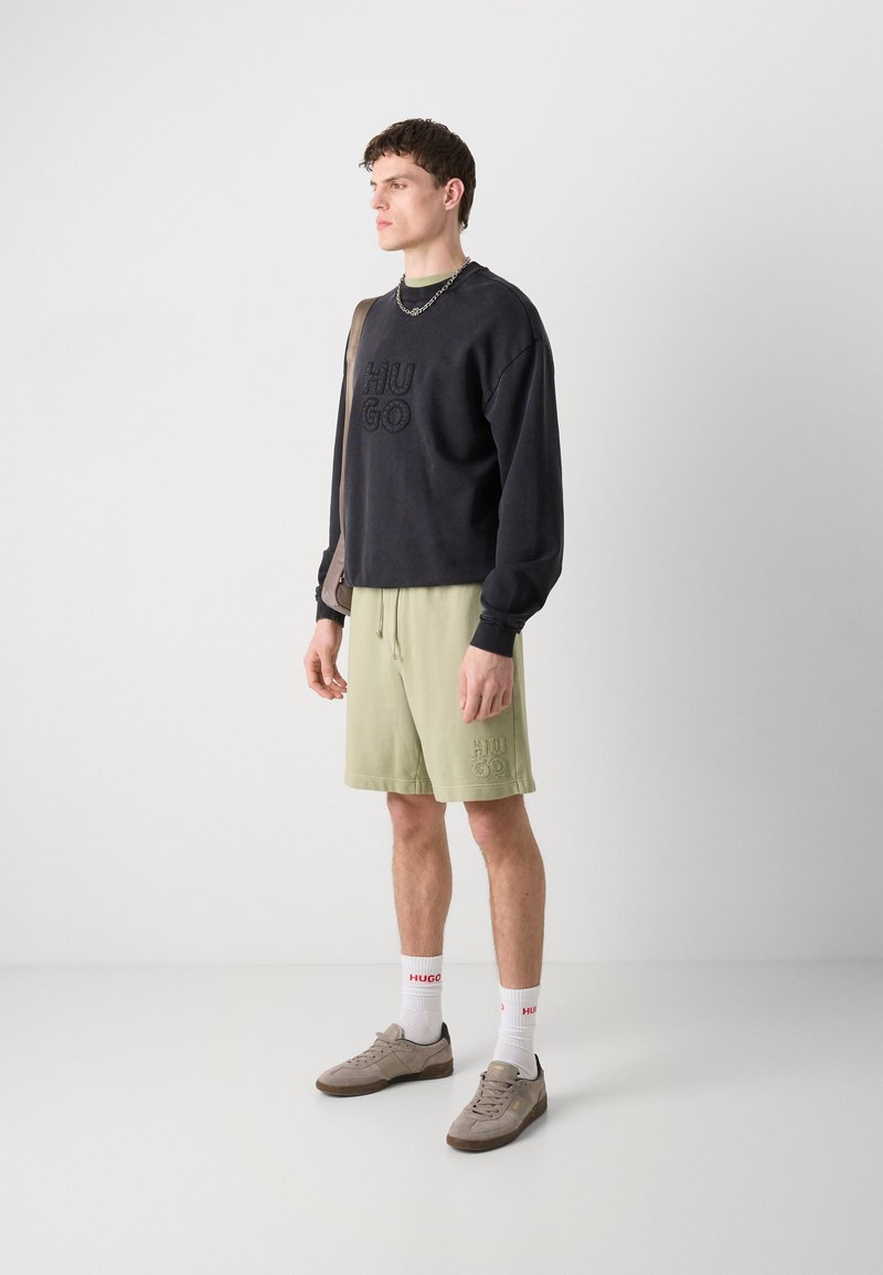 Schwarzer Sweatshirt mit "HUGO"-Logo, kombiniert mit hellgrünen Shorts mit passendem Logo. Graue Schuhe und weiße Socken mit roter Schrift runden den Look ab.