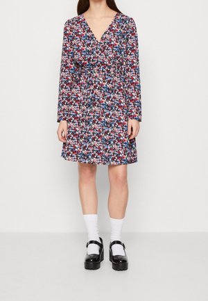 Kobieta ubrana w sukienkę w kwiaty z długim rękawem, sięgającą do kolan, w kolorach czerwonym, białym i niebieskim, białe skarpetki oraz czarne buty typu Mary Jane.