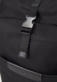 Mochila de tela negra con una textura suave, que presenta un hebilla y una correa negra prominentes, complementadas por un detalle de cuero con la marca "JOOP!".