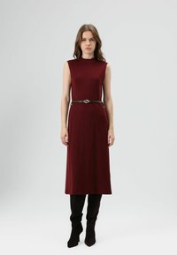 Bordeauxrode mouwloze jurk met een hoge halslijn, een getailleerde taille en een gladde textuur, gecombineerd met zwarte laarzen met hak tot aan de knie.