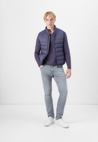 Gilet matelassé marine à motif horizontal, porté sur un pull bleu foncé. Un jean slim bleu clair et des baskets blanches complètent le look.