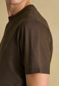 Tuvumā redzams cilvēks, kuram mugurā ir vienkārša tumši brūna t-kreklu ar īsām piedurknēm, un uz pleca un piedurknēm ir redzami šuvi sērija uz neitrāla fona.