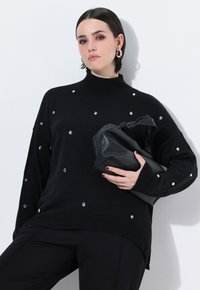 Maglione nero con collo alto, caratterizzato da dettagli decorativi in cristallo. Il modello tiene in mano una pochette in pelle nera, con texture eleganti e una vestibilità rilassata.