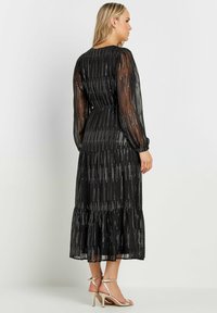 Svart långärmad klänning i genomskinligt tyg, med metalliska vertikala ränder, volangkjol och en figursydd midja. Silverfärgade klackar med remmar kompletterar.