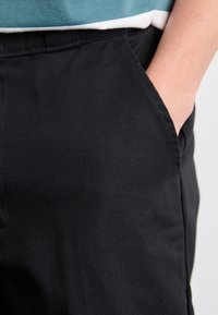 Black cotton pants with a smooth texture, featuring side pockets and a relaxed fit, shown from the waist down with a hand in the pocket.