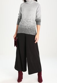 Kobieta ubrana w szary sweter z golfem z gradientem, szerokie czarne spodnie, bordowe rękawiczki w lewej ręce i bordowe botki na obcasie, stojąca.