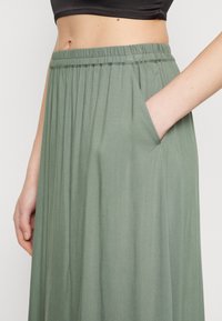Jupe plissée verte en tissu doux, dotée d'une taille élastique et d'une poche latérale, avec un design fluide et léger.