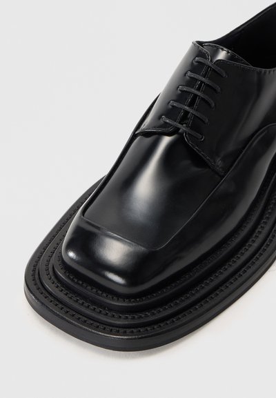 Černé leštěné kožené společenské boty s hranatou špičkou, šněrováním a prošívanou podešví na bílém pozadí.