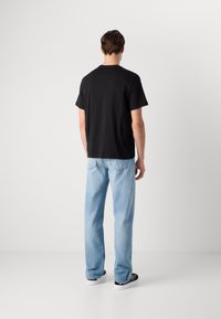 Man standing with back visible, wearing a black short-sleeve t-shirt, light blue jeans, and black and white sneakers on a plain background.