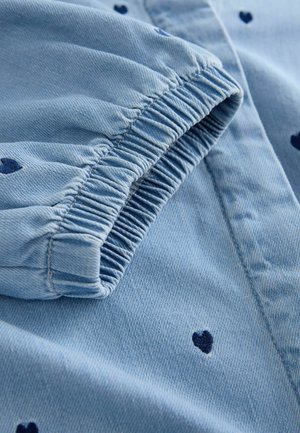 Jasnoniebieska tkanina jeansowa z ciemnoniebieskimi wzorami w kształcie serca. Elastyczny mankiet rękawa z fakturowanymi detalami szwów. Widok z bliska.