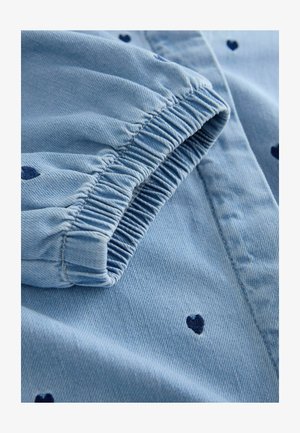 Jasnoniebieska tkanina jeansowa z ciemnoniebieskimi wzorami w kształcie serca. Elastyczny mankiet rękawa z fakturowanymi detalami szwów. Widok z bliska.