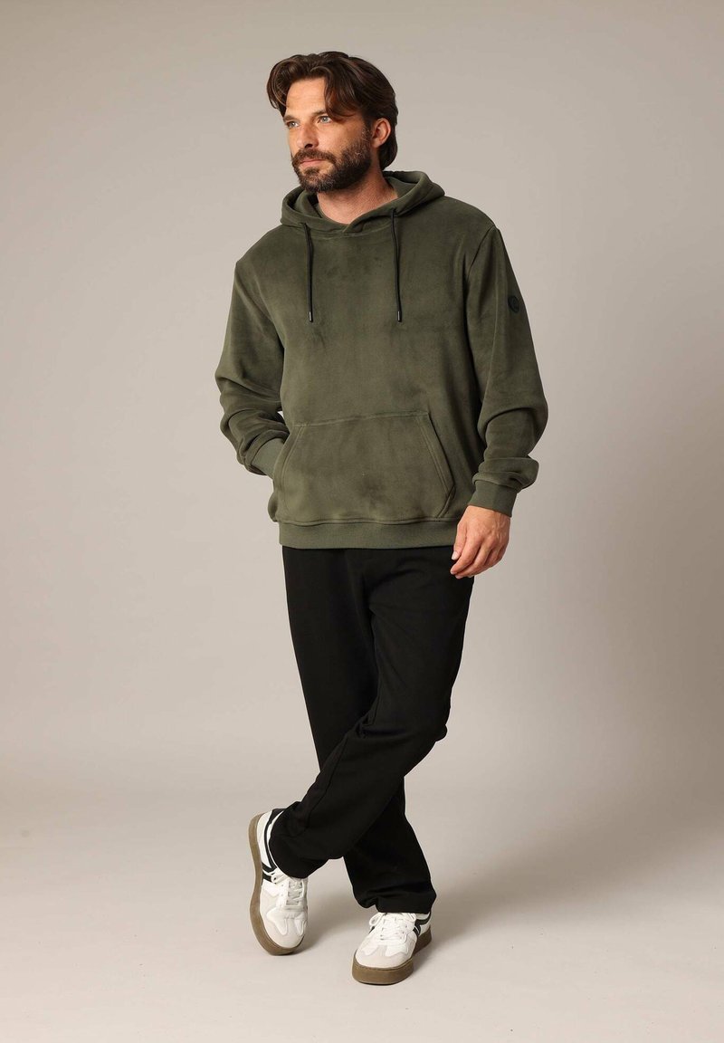 Olijfgroene fleece hoodie met een buidelzak en verstelbare trekkoorden, gecombineerd met zwarte broeken en witte sneakers, tegen een neutrale achtergrond.