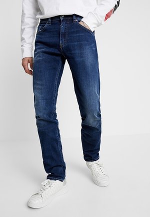 Man die een donkerblauwe spijkerbroek en witte sneakers draagt met een wit shirt met lange mouwen, staand met één hand in de zak.