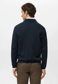 Granatowy sweter dziany z kołnierzem, długimi rękawami oraz ściągaczami na mankietach i dole. Noszony z brązowymi spodniami, prezentuje prosty design.