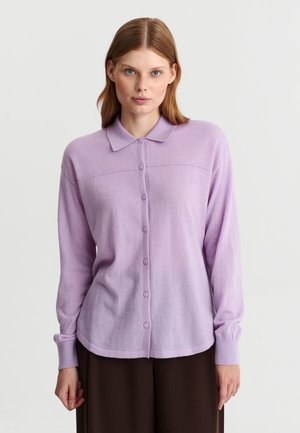 Femme portant une chemise violette claire à manches longues boutonnée avec un col et un pantalon sombre, debout devant un fond uni.