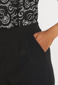 Svart spetsblus med virvlande mönster, kombinerad med mörka byxor som har en sidoficka. Slät textur och åtsittande design är synliga.