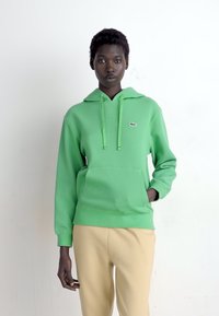 Persona di colore che indossa un hoodie verde con una tasca frontale e pantaloni beige, in piedi davanti a uno sfondo bianco uniforme.