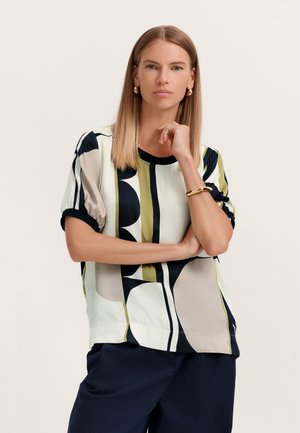 Frau mit glattem, blondem Haar, die ein kurzärmeliges Oberteil mit schwarzem, weißem, beigem und grünem geometrischem Muster sowie marineblaue Hosen trägt und mit der Hand am Kinn posiert.