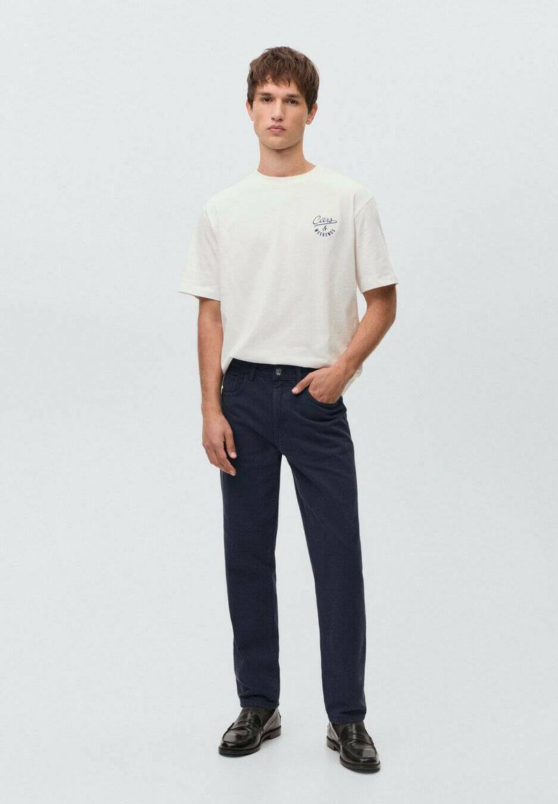 Beige bomullströja med grafiskt tryck på framsidan, matchad med marinblå byxor och svarta läderskor. En enkel, avslappnad outfit med en löst sittande passform.