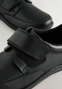 Chaussures en cuir noir avec une sangle Velcro, bout arrondi, surface lisse et coutures contrastantes. Design discret adapté à un usage décontracté.