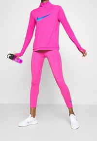 Top atletico rosa con il logo Nike blu, abbinato a leggings rosa coordinati. Tiene in mano una bottiglia d'acqua viola. Indossa sneaker bianche.