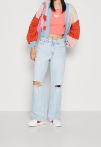 Cárdigan de punto colorido con secciones en naranja, rosa y azul; top corto rosa con corte en forma de corazón; jeans anchos rotos de color azul claro; zapatillas blancas.