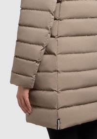 Daunenjacke in hellem Beige mit einem gesteppten Design. Sie verfügt über eine glatte Textur, lange Ärmel und ein Markenetikett auf der linken unteren Seite.