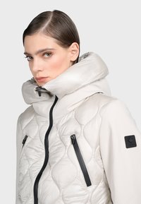 Padded kapucňová bunda v svetlej béžovej farbe s prešitým vzorom, čiernymi zipsovými vreckami a výraznou nášivkou loga na ľavom rukáve.