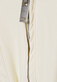 Jacke mit Reißverschluss aus einem glatten, cremefarbenen Stoff; ausgestattet mit einem metallischen Zipper und strukturiertem Nähen an den Seiten.