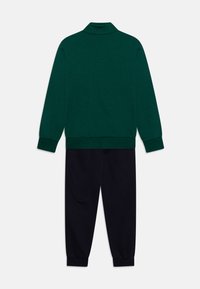 Pullover verde scuro con colletto alto e maniche lunghe, abbinato a pantaloni neri affusolati. Entrambi i capi sono realizzati in tessuto morbido.