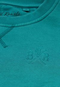 Teal-sweateri, jossa on ribbineitti ja brodeerattu "B&R" logo samalla langalla. Teksturoitu kangas hienolla neulekuviolla.