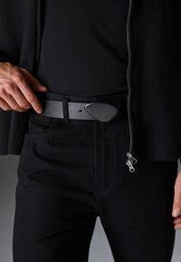 Svart läderbälte med en stor, triangulär spänne. Kombinationen med svarta byxor och en mörk topp skapar en monokromatisk look.