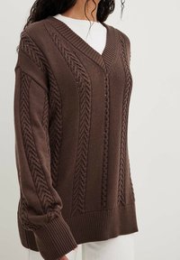 Brauner gestrickter Pullover mit V-Ausschnitt, der strukturierte Zopfmuster und gerippte Bündchen aufweist. Lockere Passform mit ausgeprägten Strickdetails.