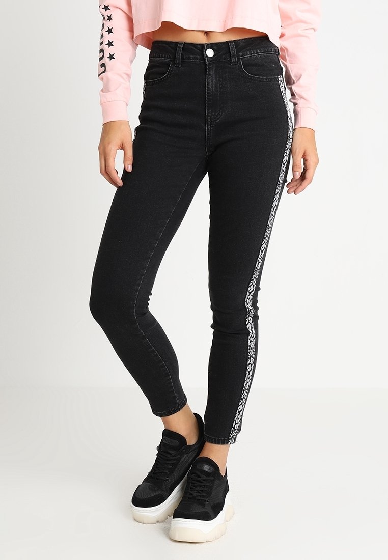 Jeans negros de cintura alta con un corte ajustado, que presentan una franja lateral blanca con un patrón. Fabricados en denim con una textura suave. Llevados con zapatillas negras.