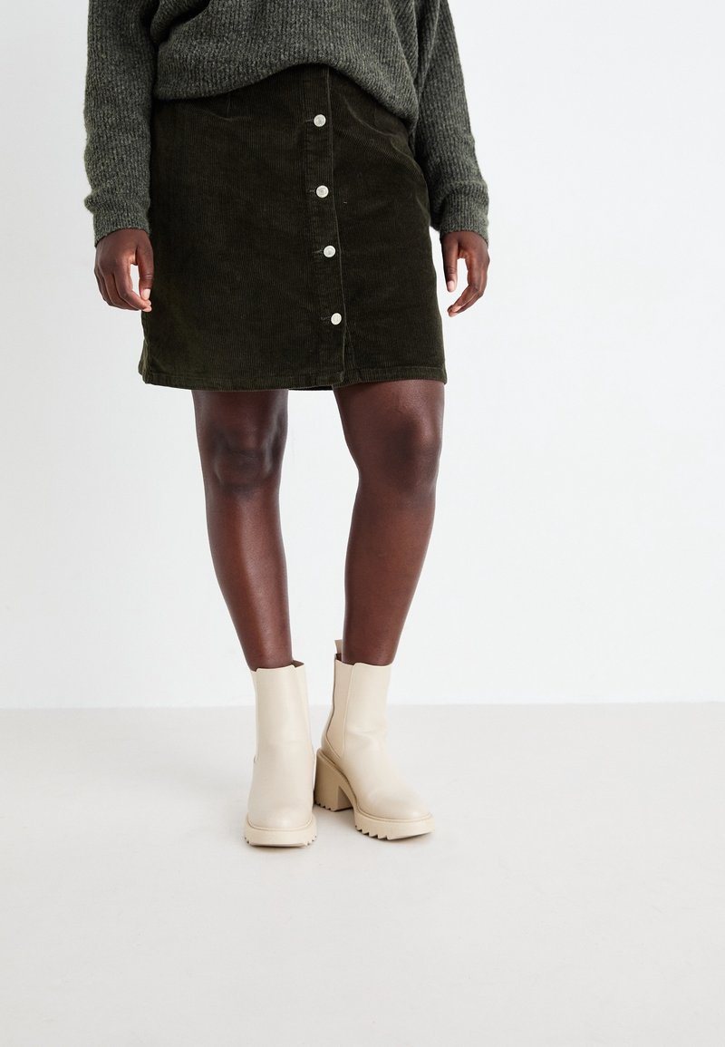 Jupe en velours côtelé vert foncé à boutons sur le devant, portée avec un pull gris et des bottines crème à talons épais sur une personne à la peau foncée.