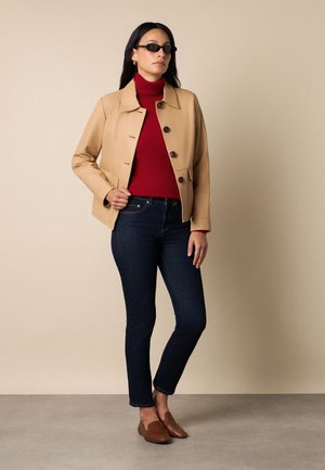 Vrouw met zwarte zonnebril, beige jasje met knopen, rode coltrui, donkerblauwe spijkerbroek en bruine loafers, staand tegen een beige muur.