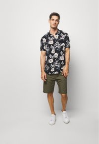 Chemise noire à manches courtes avec des fleurs blanches et roses, associée à un short vert olive et des baskets blanches, se tenant devant un fond uni.