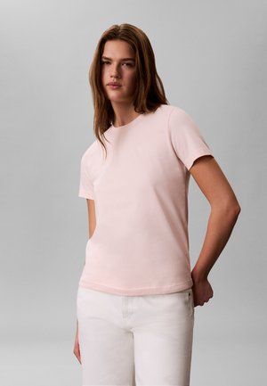 Γυναίκα που φοράει ένα ανοιχτό ροζ κοντομάνικο πουκάμισο και λευκό παντελόνι, στέκεται μπροστά σε ένα απλό γκρι φόντο.