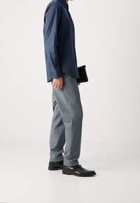 Muž oblečený v tmavě modré košili, šedých texturovaných kalhotách, černých kožených botách, držící malou černou kabelku na jednobarevném bílém pozadí.