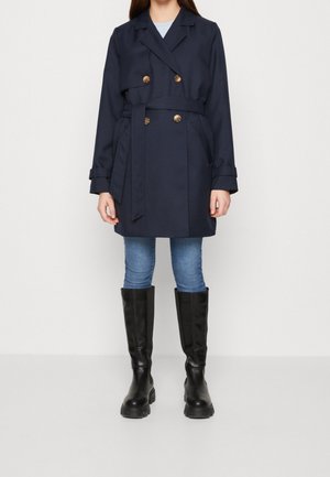 Frau trägt einen marineblauen, zweireihigen Trenchcoat mit Gürtel, hellblaue Jeans und schwarze kniehohe Stiefel und steht vor einem schlichten Hintergrund.