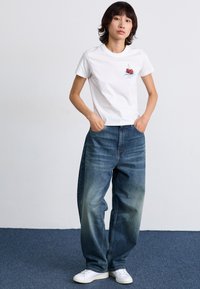 Hvid bomulds t-shirt med kirsebærgrafik, parret med løse blå jeans med en afbleget vask og brede ben, kompletteret af hvide sneakers.
