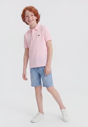 Rødhåret dreng smilende, iført en lyserød polo-shirt, blå denimbukser og hvide sneakers, stående på ét ben mod en ensfarvet baggrund.